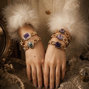 Può includere: Braccialetti a catena dorati adornati con grandi pietre preziose colorate. I braccialetti presentano pietre rettangolari e ovali nelle tonalità del viola, verde e grigio. I gioielli sono esposti sui polsi di una persona.