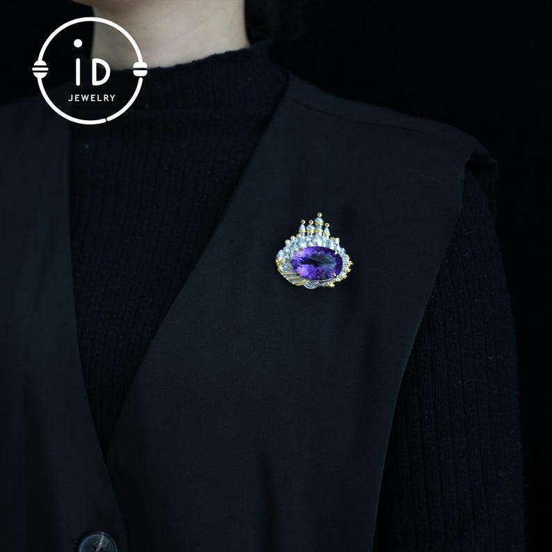 May include: A gold-toned brooch with a large, oval, purple gemstone at its center. The brooch has a crown-like design with smaller clear stones and a detailed, textured border. The brooch is pinned to a black garment.