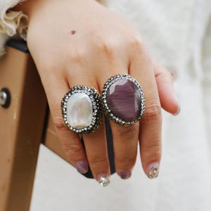 May include: Two large, ornate rings. One ring features a large, oval, pearlescent stone surrounded by small, clear crystals. The other ring has a large, oval, purple-grey stone, also encircled by small, clear crystals. Both rings have a dark metal band.
