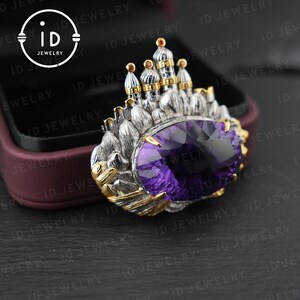 May include: A silver and gold-toned brooch featuring a large, oval-cut purple gemstone. The brooch is designed with a castle-like structure on top, with intricate detailing. The ID Jewelry logo is visible in the upper left corner.