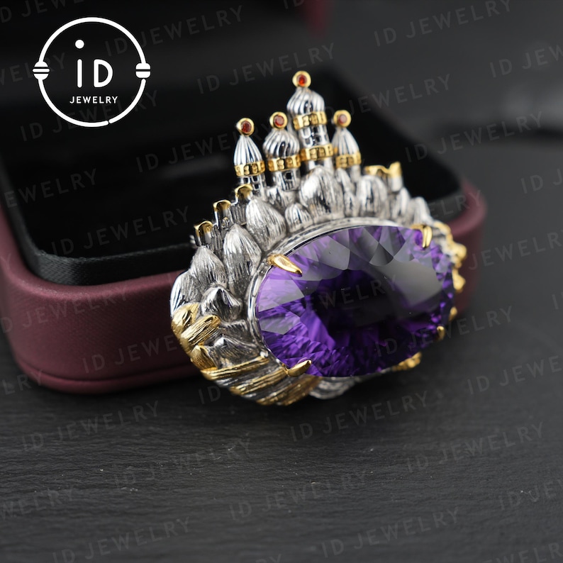 May include: A silver and gold-toned brooch featuring a large, oval-cut purple gemstone. The brooch is designed with a castle-like structure on top, with intricate detailing. The ID Jewelry logo is visible in the upper left corner.