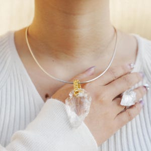 Op de afbeelding: Een ketting met een hanger in de vorm van een pijlspunt van helder kwarts met een goudkleurige beugel, gecombineerd met een bijpassende kwartsring. De ketting heeft een zilveren ketting. De sieraden worden gepresenteerd op een witte top.
