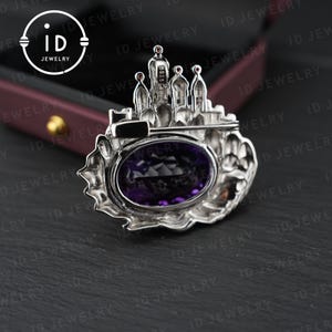 May include: A silver-toned brooch featuring a castle design with a large, oval, deep purple gemstone at its center. The brooch has intricate architectural details and small red accents. The ID Jewelry logo is visible in the background.