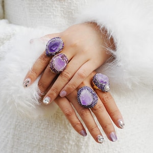 Könnte beinhalten: Fünf Amethyst-Edelsteinringe in verschiedenen Designs. Die Ringe zeigen große, unregelmäßige violette Steine, umgeben von kleineren violetten Edelsteinen und goldfarbenen Fassungen. Die Ringe werden auf einer Hand mit manikürten Nägeln präsentiert.