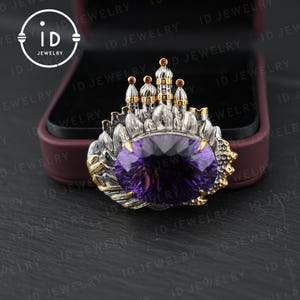 May include: A silver and gold-toned brooch featuring a large, oval-cut purple gemstone. The brooch has a castle-like design with small red accents and is set against a dark background.
