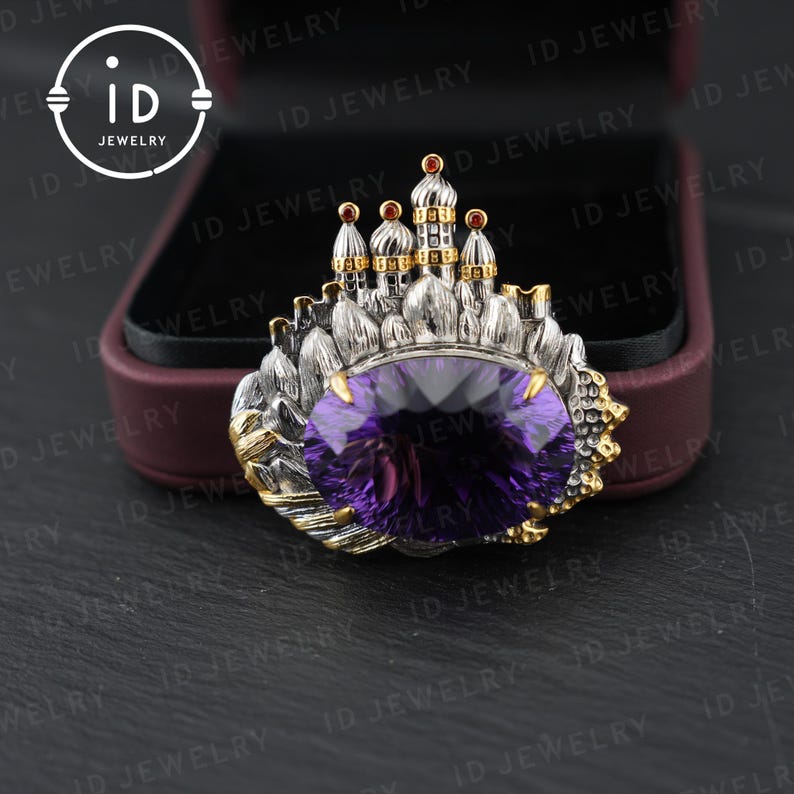 May include: A silver and gold-toned brooch featuring a large, oval-cut purple gemstone. The brooch has a castle-like design with small red accents and is set against a dark background.