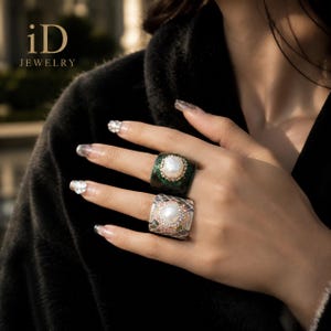May include: Two wide rings featuring central white pearls surrounded by small stones. The top ring has a dark green textured band, while the bottom ring has a grey and white patterned band. The text iD JEWELRY is visible in the upper left corner.
