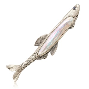 Puede incluir: Broche plateado con forma de pez y centro nacarado e iridiscente. El broche presenta escamas, aletas y cola detalladas. Una pequeña gema azul adorna el ojo. El diseño es elegante y recuerda la vida marina.