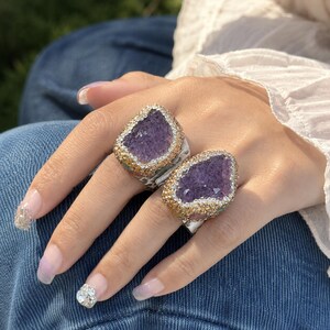 May include: Two statement rings featuring large, irregular purple druzy amethyst stones. Each stone is framed by a border of small, sparkling gold and clear crystals set in a metallic base. The rings are worn on adjacent fingers, showcasing the textured, crystalline surfaces of the deep violet gems and the reflective, multi-faceted edges of the surrounding decorative stone settings.