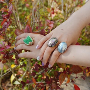 Könnte beinhalten: Drei verzierte Ringe an einer Hand. Ein Ring hat einen grünen, blattförmigen Stein mit goldenen Akzenten. Ein anderer hat einen ovalen, irisierenden Stein mit einem schwarzen Band. Der dritte Ring hat einen hellblauen, ovalen Stein mit einem silbernen Band.