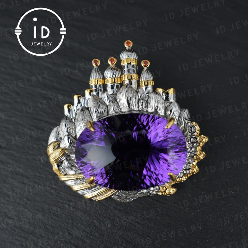 May include: A silver and gold-toned brooch featuring a large, oval-cut purple gemstone. The brooch is designed with a castle motif, with small towers and red accents. The ID Jewelry logo is in the upper left corner.