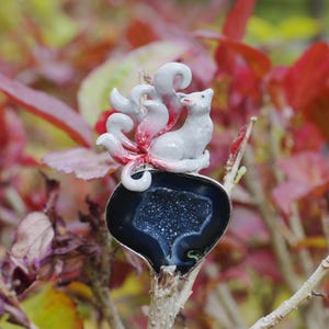 Op de afbeelding: Een broche met een gebeeldhouwde witte vos met meerdere staarten met rode punten, zittend op een donkere, druppelvormige geode-schijf. De geode heeft een gepolijste zwarte buitenrand en een sprankelend, diepblauw kristallijn centrum met witte spikkels.