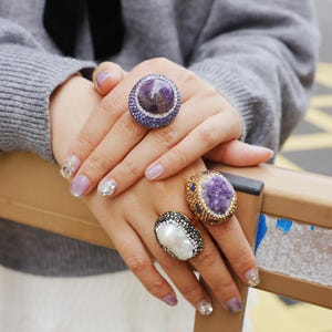 Può includere: Primo piano di anelli con pietre preziose di grandi dimensioni. Un anello presenta una grande pietra preziosa rotonda viola circondata da pietre viola più piccole. Un altro anello ha una perla bianca con dettagli neri e argento. Un terzo anello ha una montatura dorata con un cristallo viola.