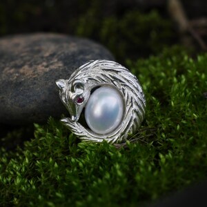 Puede incluir: Un broche de plata con forma de zorro enroscado con detalles de pelaje texturizado. El zorro presenta una pequeña piedra preciosa roja y redonda como ojo y rodea una perla blanca, lisa y ovalada en su centro, creando un diseño circular.