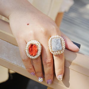 May include: Two ornate rings with gold-colored bands are displayed. One ring features a red, oval-shaped center stone, while the other has a rectangular, light gray stone. Both are surrounded by small, white pearls.