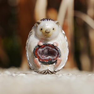 Peut inclure: Une figurine de hérisson fantaisiste avec un corps blanc, des détails argentés et un ventre en géode. La géode présente des nuances de rouge et de violet. Le hérisson a de petits pieds roses et une tête brune.
