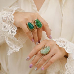 Op de afbeelding: Drie sierlijke ringen met grote, kleurrijke edelstenen. Eén ring heeft een diepgroene ovale steen, een andere een gebeeldhouwde groene jade en de derde een lichtgroene ovale steen. Alle zijn gezet in goudkleurig metaal met kleinere stenen.