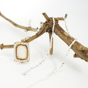 Puede incluir: Un collar con colgante rectangular con una piedra central blanca, enmarcada por pequeñas perlas y cuentas doradas. El colgante cuelga de una delicada cadena plateada. El collar se exhibe sobre una pequeña rama de madera natural sobre un fondo blanco.