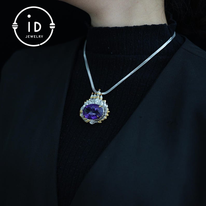 May include: A silver necklace with a large, oval, purple gemstone pendant. The pendant is framed with gold and silver accents. The necklace is displayed against a black background. The pendant is a statement piece of jewelry.
