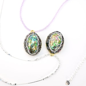 Puede incluir: Dos colgantes ovalados con conchas de abulón iridiscentes. Cada concha está bordeada por pequeñas piedras de color gris oscuro. Un colgante cuelga de un collar de cuentas moradas claras, el otro de una cadena plateada. El fondo es blanco.