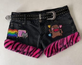 May include: Black denim shorts featuring a pixelated cat with a rainbow trail on the left and a brown character with hearts on the right. The leg openings are trimmed with pink and black faux fur. A black studded belt with a silver buckle is included.