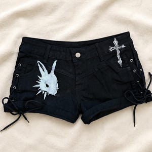 May include: Black denim shorts featuring side lace-up details with black cords and silver-tone grommets. The front displays a white graphic of a rabbit head with spikes and a white graphic of a cross on the right side. The shorts have a button closure.