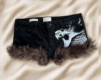 May include: Black denim shorts featuring a white graphic print of a snake head on the right side. The hem of the shorts is trimmed with long, dark brown faux fur. The shorts include a standard button closure, belt loops, and two front pockets.