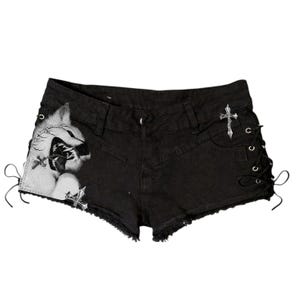 May include: Black denim shorts featuring a frayed hem, side lace-up detailing with metal eyelets, and a graphic print of a snarling wolf on the left leg. A silver-toned cross emblem is positioned on the right side near the waistband.