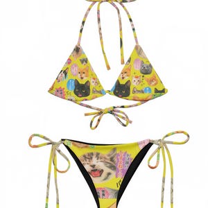 以下が含まれることがあります： 猫をテーマにしたプリントの黄色いビキニセット。トップはトライアングルカップのホルタースタイルで、ボトムはクラシックなビキニカットです。プリントには、さまざまな猫の顔と日本語のテキストが描かれています。
