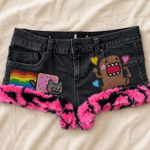 May include: Black denim shorts featuring a pixelated cat with a rainbow trail on the left side and a brown character with a wide mouth and hearts on the right. The leg openings are trimmed with a thick, textured band of bright pink and black faux fur.