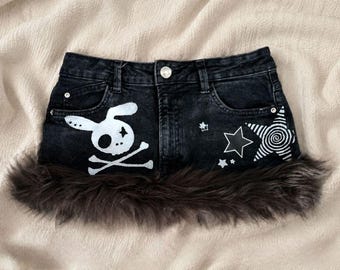 May include: A black denim mini skirt featuring a white graphic of a rabbit skull and crossbones on the left side and white star designs on the right side. The hem of the skirt is trimmed with a thick band of dark brown faux fur.