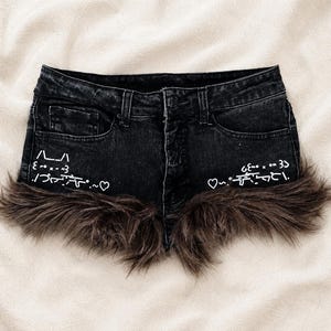 Könnte beinhalten: Schwarze Jeansshorts mit einem dunkelbraunen Kunstfellbesatz am unteren Saum. Auf der Vorderseite der Shorts befinden sich weiße, gestickte Strichgrafiken eines Katzengesichts und eines Gewehrs auf jeder Seite, ergänzt durch kleine Herzen.