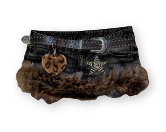 May include: A black denim mini skirt featuring a dark brown faux fur trim along the hem. The skirt includes a brown studded belt with a silver buckle, a dangling heart-shaped leopard print charm, and a white star graphic with a spiral centre.