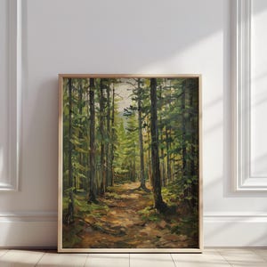 Puede incluir: Pintura enmarcada de un paisaje forestal. Altos árboles verdes bordean un camino que conduce a la distancia, con la luz del sol filtrándose a través del dosel. La obra de arte se exhibe sobre un suelo de madera contra una pared blanca.
