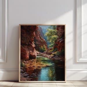 Puede incluir: Una pintura de paisaje enmarcada representa una escena de cañón con formaciones rocosas rojas y un río claro. La obra de arte presenta una representación detallada del entorno natural, con la luz del sol iluminando el agua y el follaje. La pintura se exhibe sobre un suelo de madera.