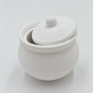Könnte beinhalten: Eine kleine, weiße Zuckerdose aus Keramik mit passendem Deckel. Der Deckel hat einen kleinen, runden Knopf oben. Die Schüssel ist rund mit einem leicht ausgestellten Rand. Der Deckel ist teilweise geöffnet und gibt den Blick ins Innere frei.