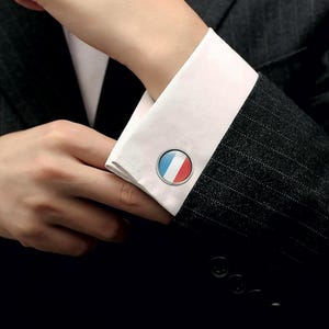 Puede incluir: Gemelos plateados redondos con el diseño de la bandera francesa. Los gemelos tienen un esquema de color azul, blanco y rojo. Están en un puño de camisa blanca, y la persona lleva un traje de raya diplomática gris oscuro.
