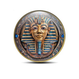 May include: A circular, gold-coloured decorative item depicting the bust of Tutankhamun. The pharaoh's face is gold with blue and gold striped headdress. The outer ring has hieroglyphs and blue accents.