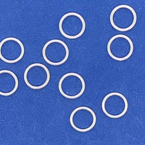 May include: Ten silver jump rings on a blue background. The jump rings are all the same size and are arranged in a random pattern.