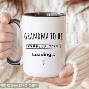May include: White ceramic mug with black interior and handle, featuring the text "Grandma to Be" above a loading bar with hearts and the year 2026. Below, the word "Loading..." and a heart symbol are displayed.