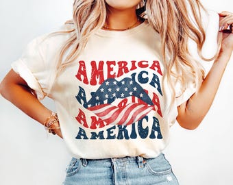 May include: Cream-colored t-shirt with the word "AMERICA" in red and blue, with a graphic of lips in the colors of the American flag. The shirt has rolled-up sleeves and is worn with blue jeans.