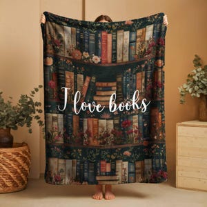 May include: A dark green throw blanket featuring a bookshelf design with various books and floral accents. The blanket has the text "I love books" in white script. The blanket is held up by a person.