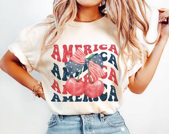 May include: Cream-colored t-shirt with the word "AMERICA" in red and blue, featuring a cherry design with a bow and stars. The shirt has rolled-up sleeves and a casual fit.