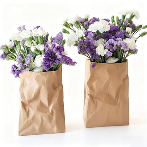 Op de afbeelding: Twee boeketten bloemen in bruine papieren zakvazen. De bloemen zijn een mix van witte anjers en paarse statice. De papieren zakken hebben een gekreukte textuur en staan tegen een witte achtergrond.