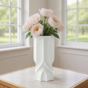 Puede incluir: Un jarrón geométrico blanco contiene un ramo de flores de color rosa pálido con centros amarillos y hojas verdes. El jarrón tiene un diseño retorcido único en la base y está sobre una mesa con tapa de mármol cerca de una ventana.