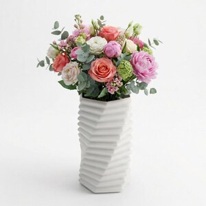 Op de afbeelding: Een levendig boeket met roze, perzik en witte bloemen, waaronder rozen en pioenrozen, in een witte geometrische vaas. De vaas heeft een zigzag ontwerp. De opstelling staat tegen een witte achtergrond.