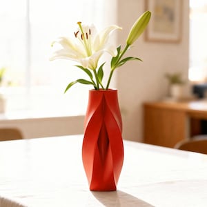 Puede incluir: Un jarrón rojo moderno con un diseño retorcido único, que contiene lirios blancos con tallos y hojas verdes. El jarrón está hecho de un material sólido y se encuentra sobre una superficie blanca. Las flores están en plena floración.