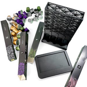 May include: An assortment of coffee pods in various colors, including gold, purple, and green, alongside black storage containers and boxes. The boxes feature botanical illustrations and the Nespresso logo. The image showcases a modern coffee setup.