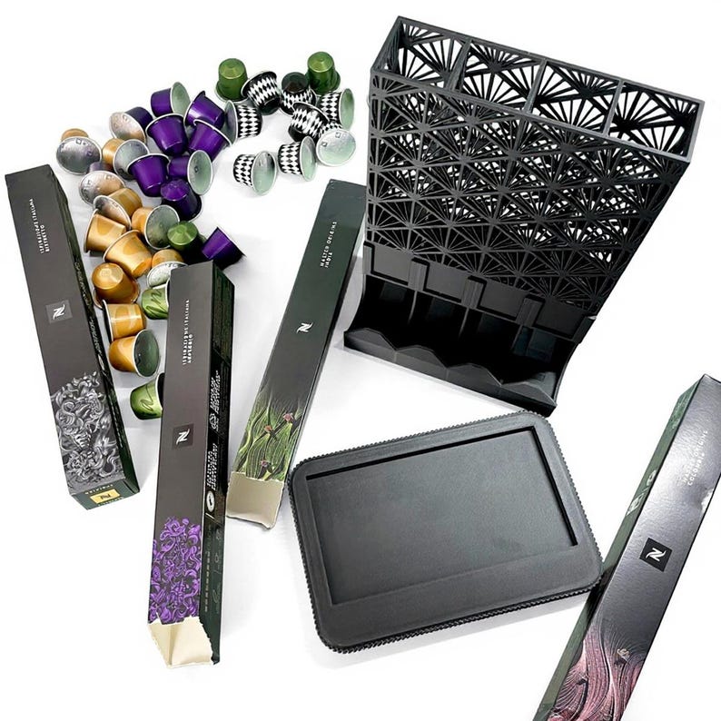 May include: An assortment of coffee pods in various colors, including gold, purple, and green, alongside black storage containers and boxes. The boxes feature botanical illustrations and the Nespresso logo. The image showcases a modern coffee setup.