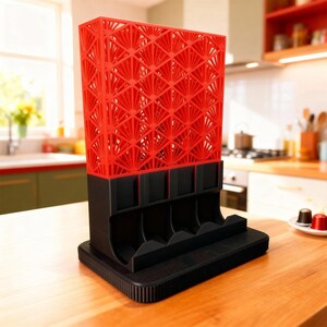 May include: A red and black coffee pod holder with a geometric fan-like design. The top section is red with a lattice pattern, and the base and pod slots are black. The holder sits on a wooden surface.